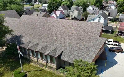 Aerial view of a large building with a gabled roof by Shane Fisk Roofing, surrounded by residential houses, cars parked nearby, and trees in the background.
