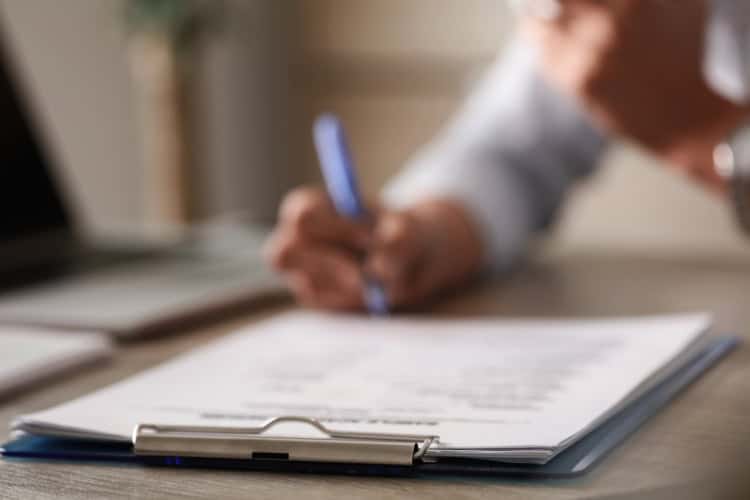 A person writes on a document attached to a clipboard, with a pen in hand, at a desk. The focus is on the clipboard and paper—perfect for recording details for Shane Fisk Roofing—with the person blurred in the background.