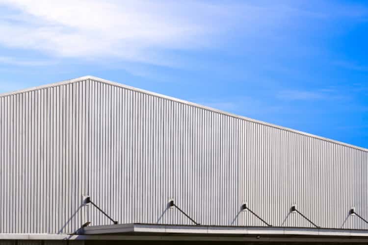 A large industrial metal building with a corrugated exterior and a sloped roof is shown against a blue sky with light clouds.