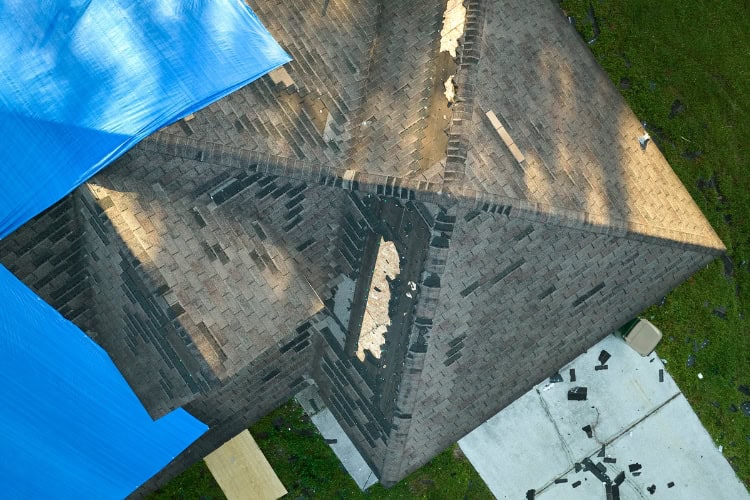 Aerial view of a house roof with missing shingles and visible damage, partially covered by a blue tarp. Debris is scattered on the ground near the house—perfect example of when to call Shane Fisk Roofing for expert repairs.