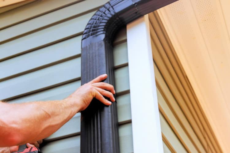 A worker installs a black downspout on the side of a house with light-colored siding and a beige soffit.