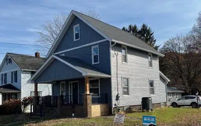 A two-story blue-gray house with a covered front porch, brown pillars, and a sloped roof by Shane Fisk Roofing sits on a grassy lot with a campaign sign in the yard.