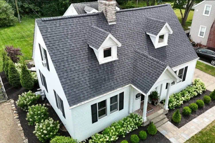 Aerial view of a white two-story house with a dark gray Shane Fisk Roofing roof, dormer windows, a small front porch, and landscaped greenery surrounding the exterior.