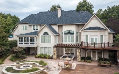 Two-story beige house with a dark roof, large windows, a wooden deck with pergola, and a landscaped backyard featuring a circular patio area.