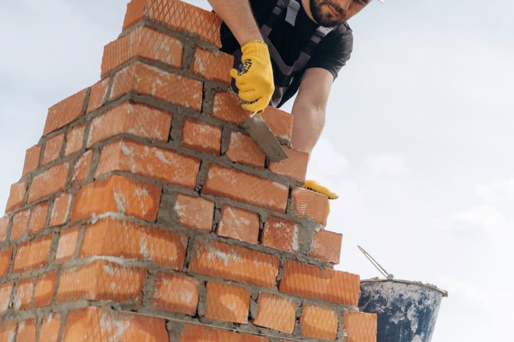 A mason, wearing gloves, uses a trowel to lay bricks and mortar on a partially built brick chimney.