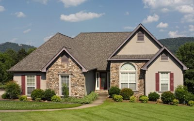 Single-story suburban house with stone and stucco exterior, gable roof, red shutters, large front lawn, and shrubs, set against a backdrop of trees and hills.
