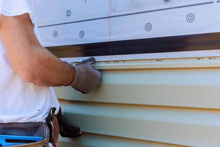 A person wearing gloves installs light green horizontal vinyl siding on a house exterior using a measuring tool to ensure perfect alignment.