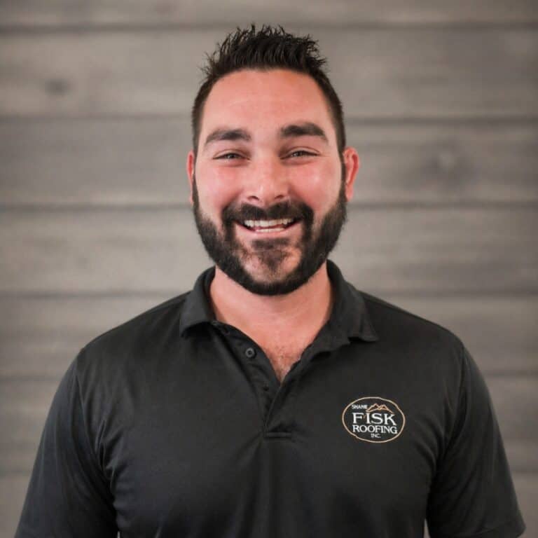 A man with dark hair and a beard smiles at the camera, wearing a black Shane Fisk Roofing polo shirt, standing in front of a wooden wall background.