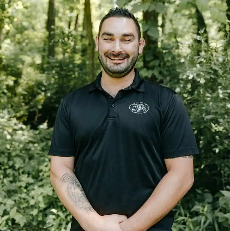 A man with dark hair and a beard, wearing a black polo shirt with the Shane Fisk Roofing logo, stands smiling outdoors in a wooded area with greenery in the background.