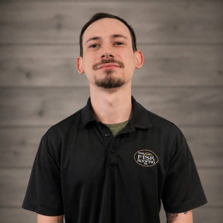 A man with short dark hair and a goatee wears a black Shane Fisk Roofing polo shirt, standing in front of a gray wooden wall.