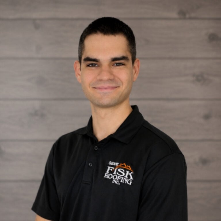 A man with short dark hair wearing a black Shane Fisk Roofing Inc. polo shirt stands in front of a wood-paneled background, smiling at the camera, giving a glimpse into what Shane Fisk Roofing is all about.