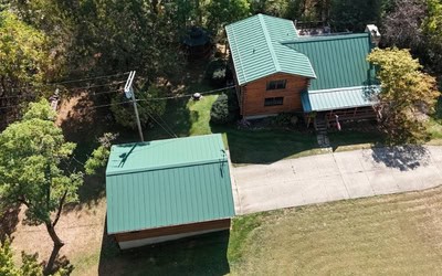 Aerial view of a house with a green metal roof installed by Shane Fisk Roofing, a detached garage, concrete driveway, and surrounding trees and lawn.