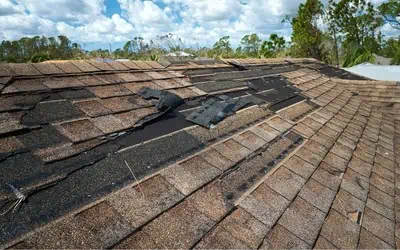 A shingled roof with several damaged and missing shingles, exposing black underlayment beneath, sits beneath a partly cloudy sky—an issue the experts at Shane Fisk Roofing can repair efficiently.