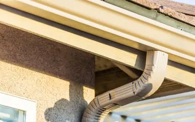 Close-up of a beige rain gutter and downspout attached to the roof edge of a house, with sunlight casting shadows on the stucco exterior wall.
