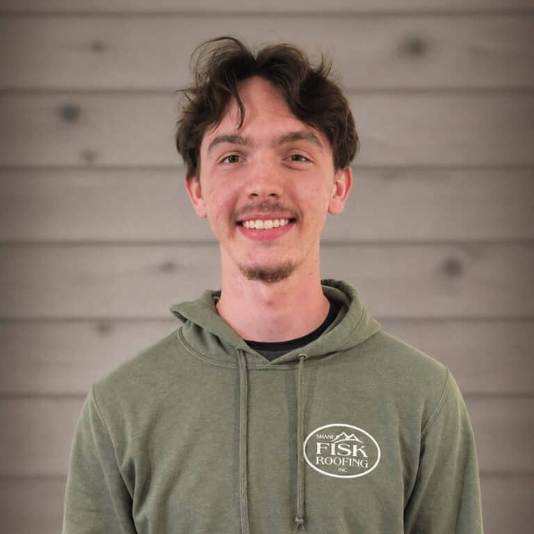Young man with brown hair and a goatee wearing a green hooded sweatshirt with a "Shane Fisk Roofing" logo, standing in front of a wooden plank wall, smiling at the camera.