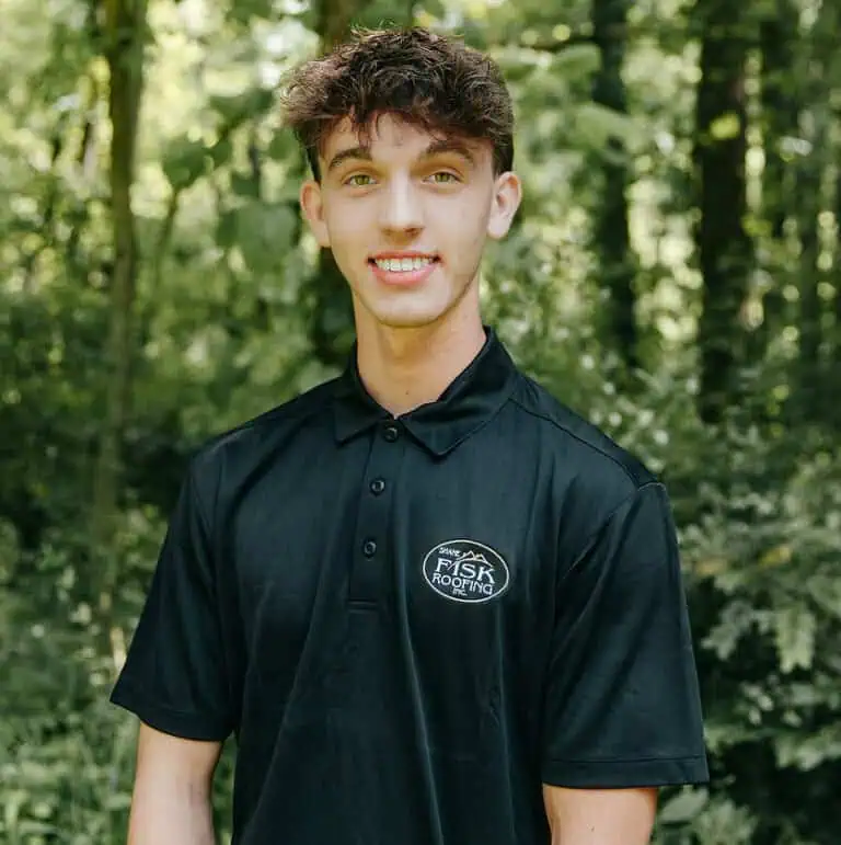 Young man stands outdoors in front of green trees, wearing a black polo shirt with a "Shane Fisk Roofing" logo on the chest, smiling at the camera.