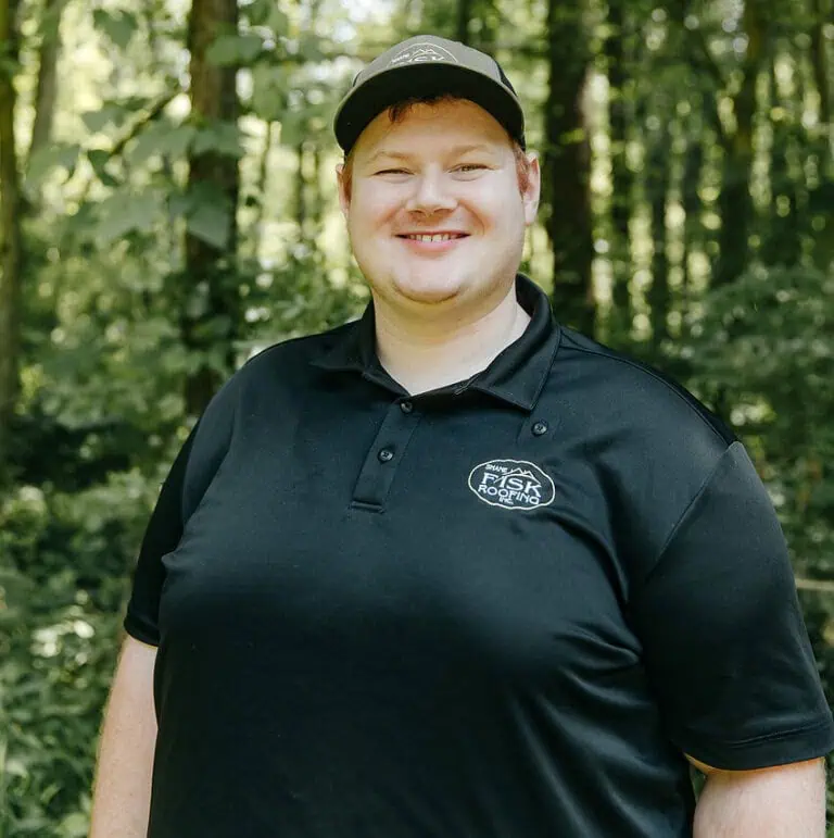 A man wearing a black polo shirt and cap stands outdoors in a wooded area, smiling at the camera—representing Shane Fisk Roofing.