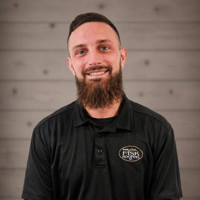 A man with a beard in a black "Shane Fisk Roofing" polo shirt stands in front of a gray wooden panel wall, smiling at the camera.