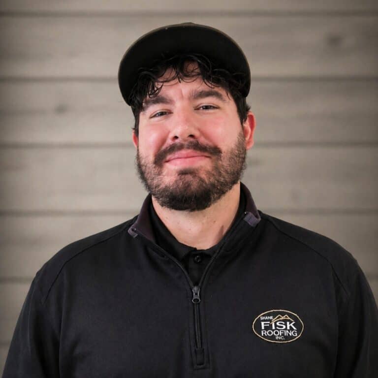 A man with a beard and dark hair wearing a black cap and a black shirt with a "Shane Fisk Roofing" logo stands in front of a wooden wall.