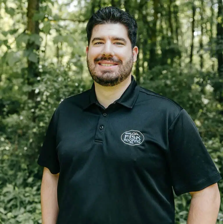 A man with dark hair and a beard stands outdoors in front of trees, wearing a black polo shirt with a "Shane Fisk Roofing" logo on the chest.