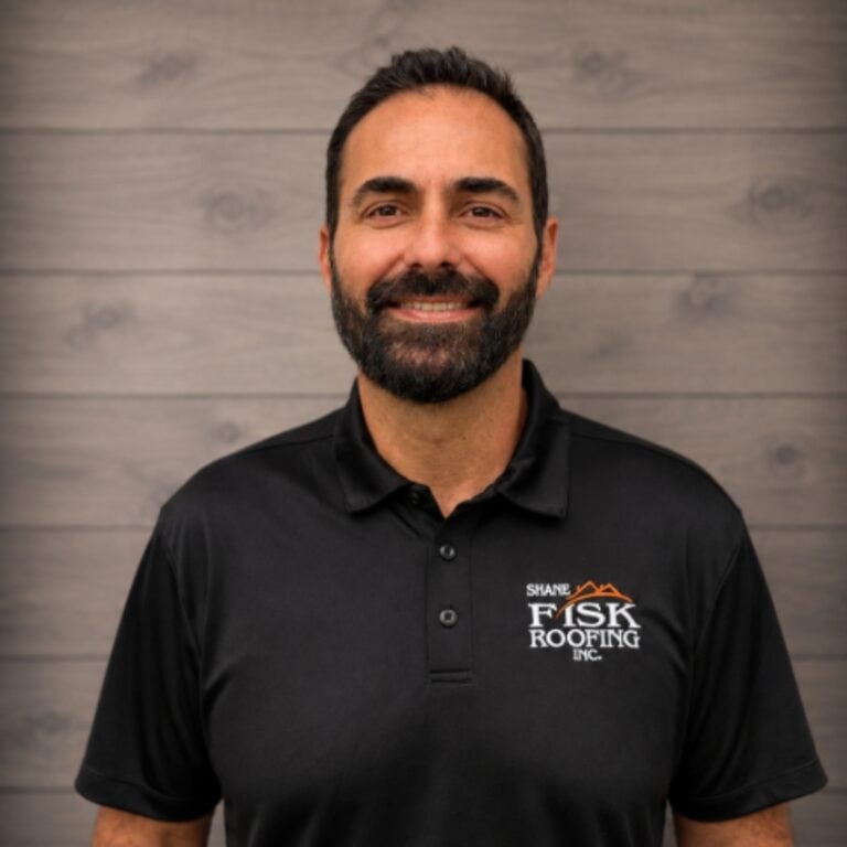 Man with dark hair and beard wearing a black polo shirt with the “Fisk Roofing Inc.” logo, standing in front of a wood panel background—representing Shane Fisk Roofing’s commitment to quality and professionalism.