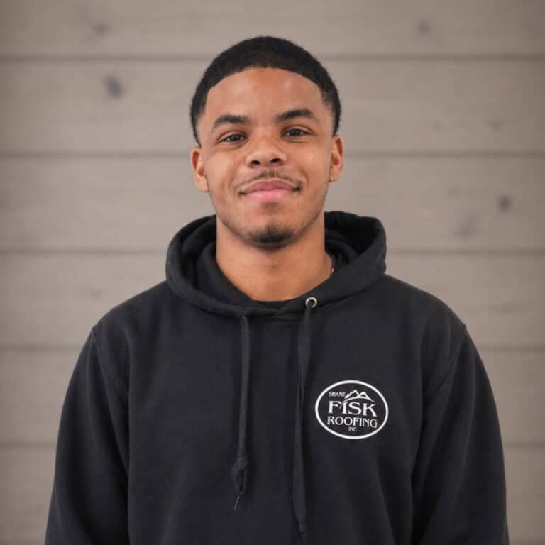 A man stands in front of a wooden wall, wearing a black hoodie with a "Shane Fisk Roofing" logo on the chest, and smiles slightly at the camera.