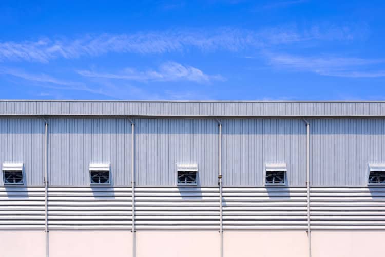Exterior view of a large industrial building featuring metal wall panels with small ventilation windows under a blue sky with scattered clouds.