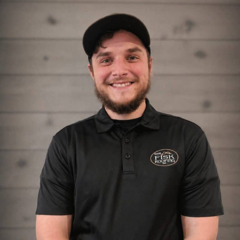 A man wearing a black Shane Fisk Roofing polo shirt and a black cap stands in front of a gray wooden wall, smiling at the camera.