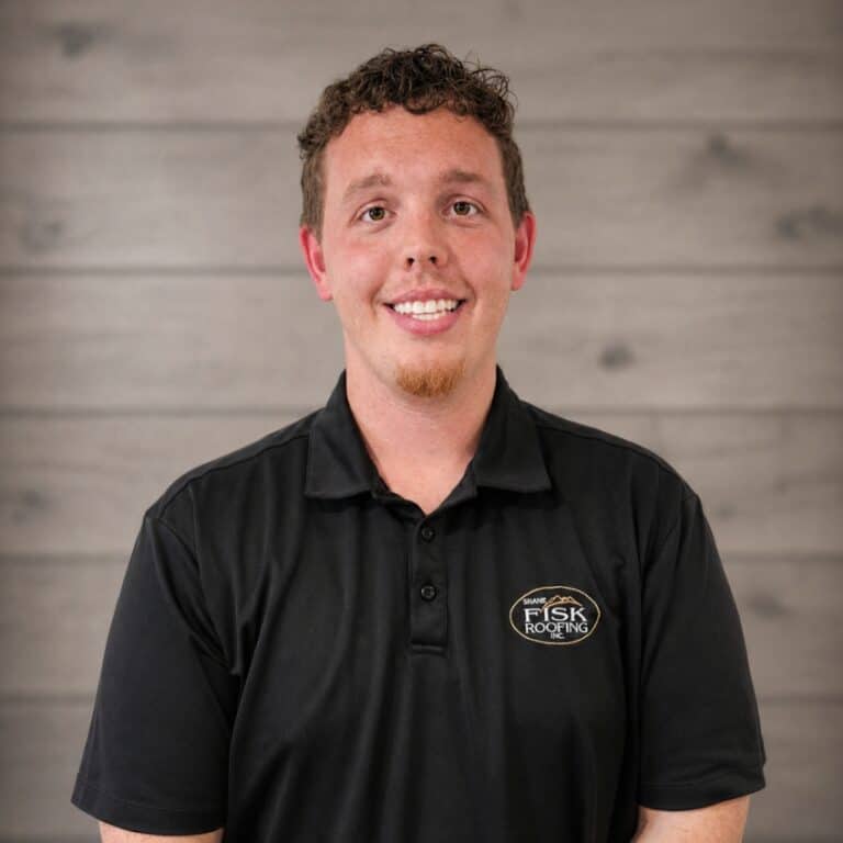 A man with short curly hair wearing a black Shane Fisk Roofing polo shirt stands in front of a gray wooden wall, smiling at the camera.