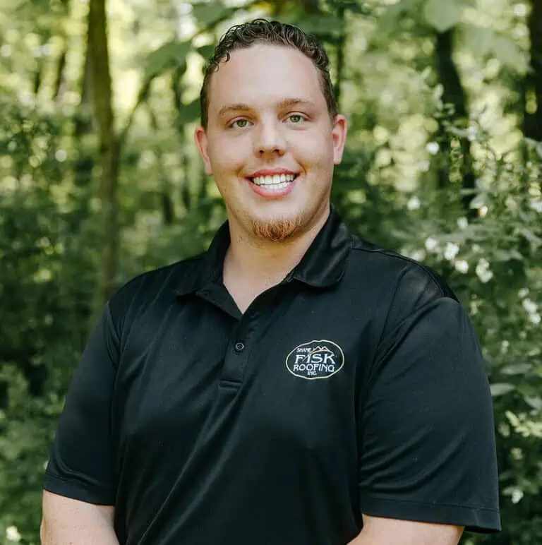 A man in a black polo shirt with a "Shane Fisk Roofing" logo stands outdoors in front of green foliage, smiling at the camera—proudly representing Shane Fisk Roofing.