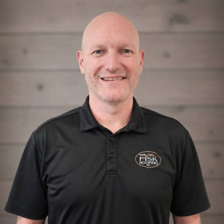 A man wearing a black Shane Fisk Roofing polo shirt stands smiling in front of a gray wooden wall.