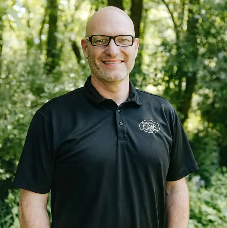 Bald man with glasses wearing a black polo shirt stands outdoors in front of green trees, smiling at the camera, representing Shane Fisk Roofing.