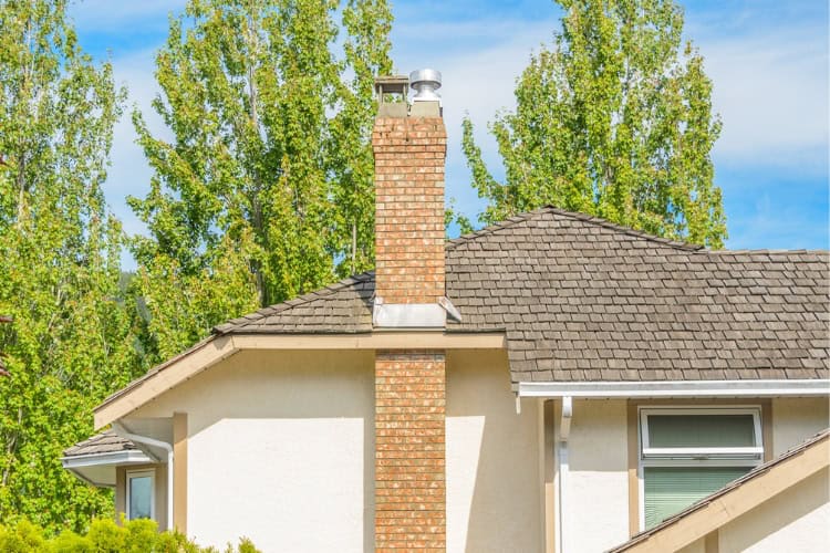 A beige house with a shingle roof and a red brick chimney, set against tall green trees and a blue sky.