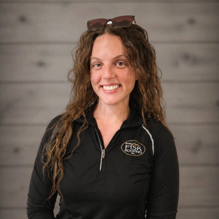 A woman with curly brown hair wearing sunglasses on her head and a Shane Fisk Roofing zip-up shirt poses in front of a wooden wall background.