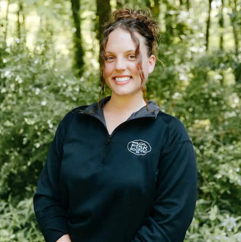 A person with curly hair smiles outdoors, wearing a black zip-up pullover with a "Shane Fisk Roofing" logo, standing in front of green foliage.