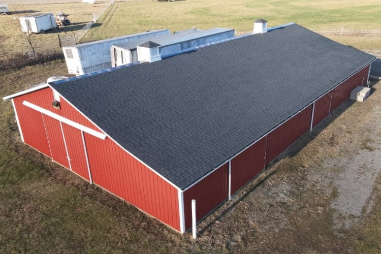 A large red metal barn with a dark shingle roof by Shane Fisk Roofing and white trim stands on a gravel area in a grassy, rural landscape.