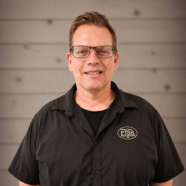 A man wearing glasses and a black "Shane Fisk Roofing" shirt stands in front of a gray wooden wall, smiling at the camera.