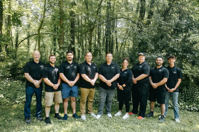 Nine people stand in a row outdoors, all wearing black Shane Fisk Roofing shirts and posing for a group photo in front of a wooded background.