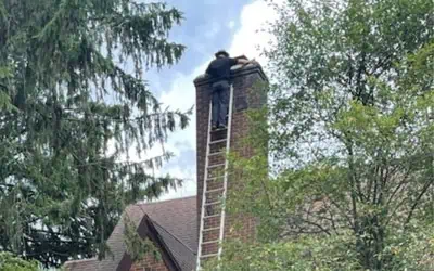 A person from Shane Fisk Roofing is standing on a ladder, working at the top of a brick chimney on a house, surrounded by trees.