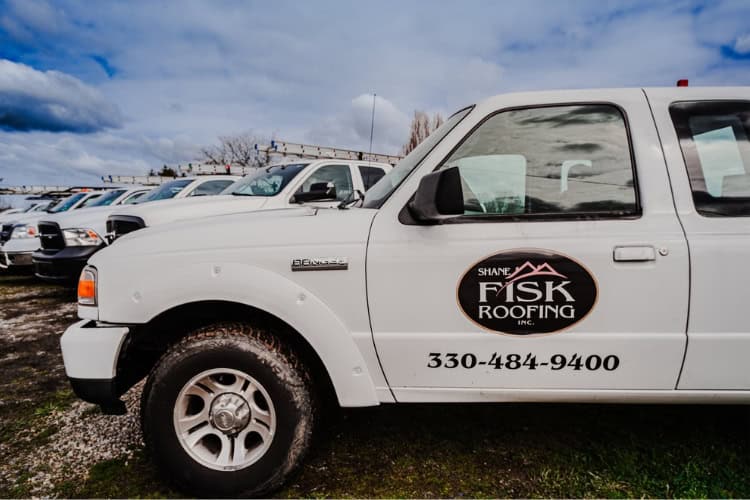 A white pickup truck with "Shane Fisk Roofing Inc." and a phone number on the door is parked in a row with other Shane Fisk Roofing vehicles.