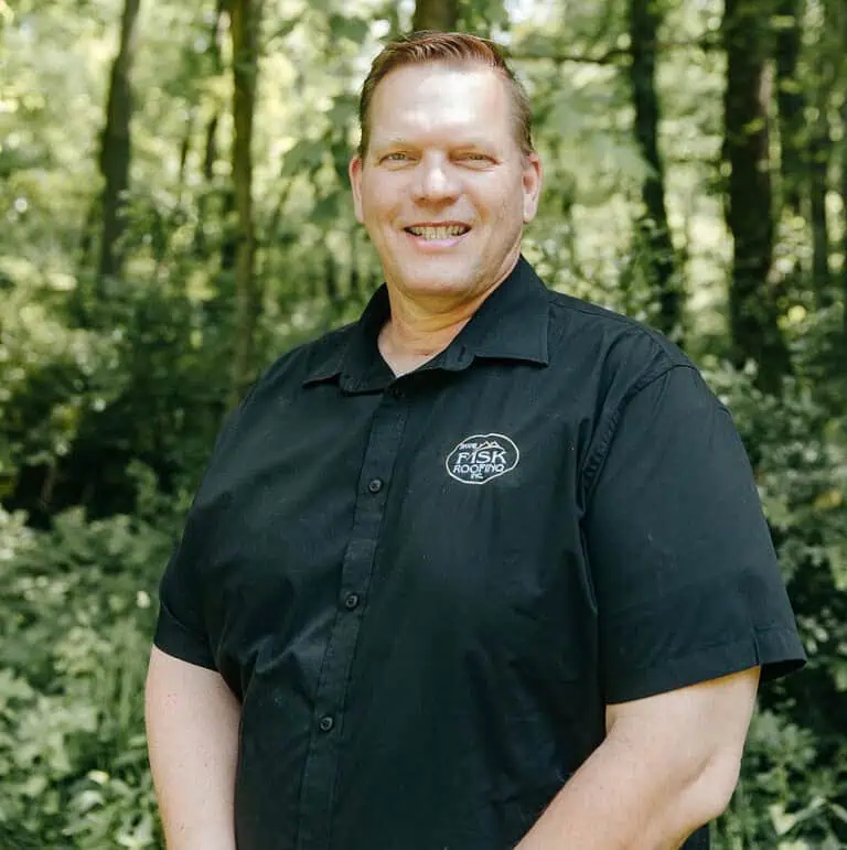 A man wearing a black short-sleeve Shane Fisk Roofing button-up shirt with a logo stands outdoors in a wooded area, smiling at the camera.