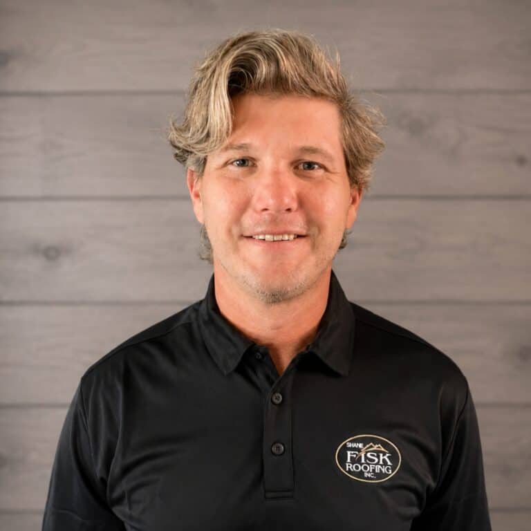 A man with wavy blonde hair wearing a black Shane Fisk Roofing polo shirt stands in front of a wood-paneled background, facing the camera and smiling.