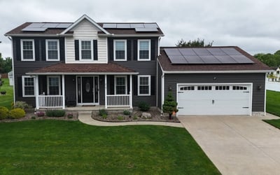 Two-story house with dark siding, white trim, front porch, attached garage, and solar panels on both the garage and main roof. This home features quality roofing throughout. The lawn is green and the driveway is concrete.