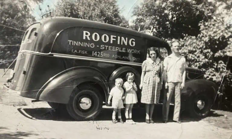 A black-and-white photo of a man, woman, and two young children standing beside a vintage Shane Fisk Roofing vehicle with “Roofing, Tinning, Steeple Work” signage on the side.