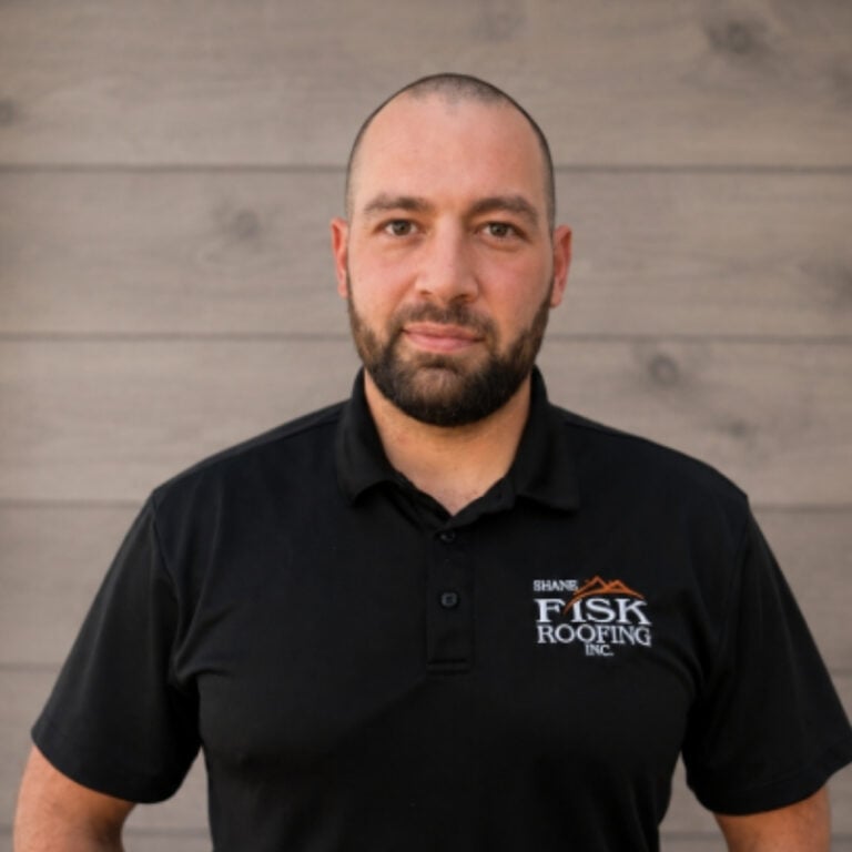 A man with a shaved head and beard wearing a black FISK Roofing polo shirt stands in front of a wooden wall, facing the camera with a neutral expression, representing the professionalism at Shane Fisk Roofing.
